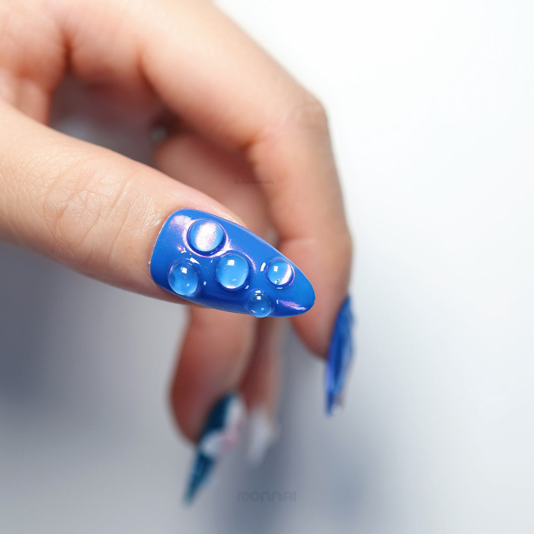 Petal Waves press-on nails in close-up, blue with glossy raised bubble accents and a soft luminous shimmer.