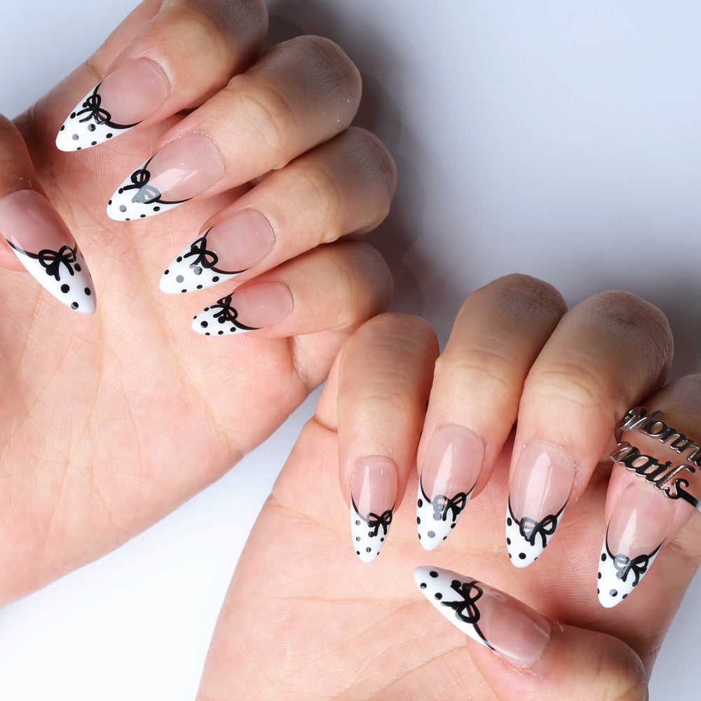 Polka Charm press-on nails worn on hands, sheer nude with white French tips, black bows and polka dots.