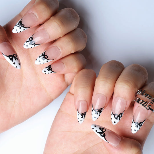 Polka Charm press-on nails worn on hands, sheer nude with white French tips, black bows and polka dots.
