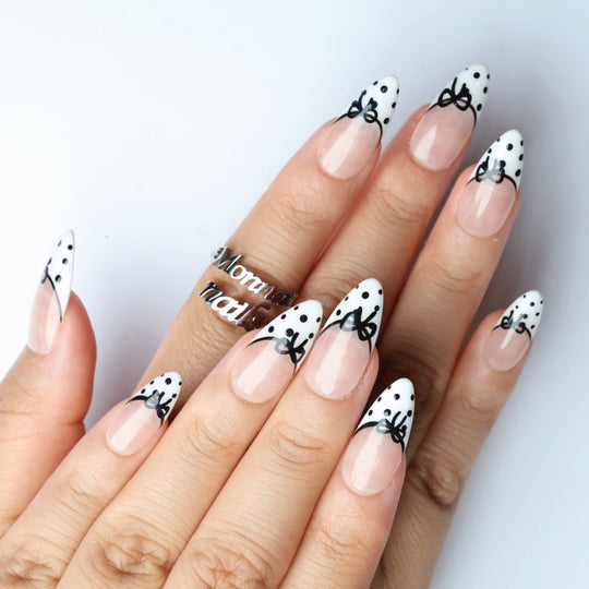 Polka Charm press-on nails worn on hands, sheer nude with black bows, white French tips and black polka dots.