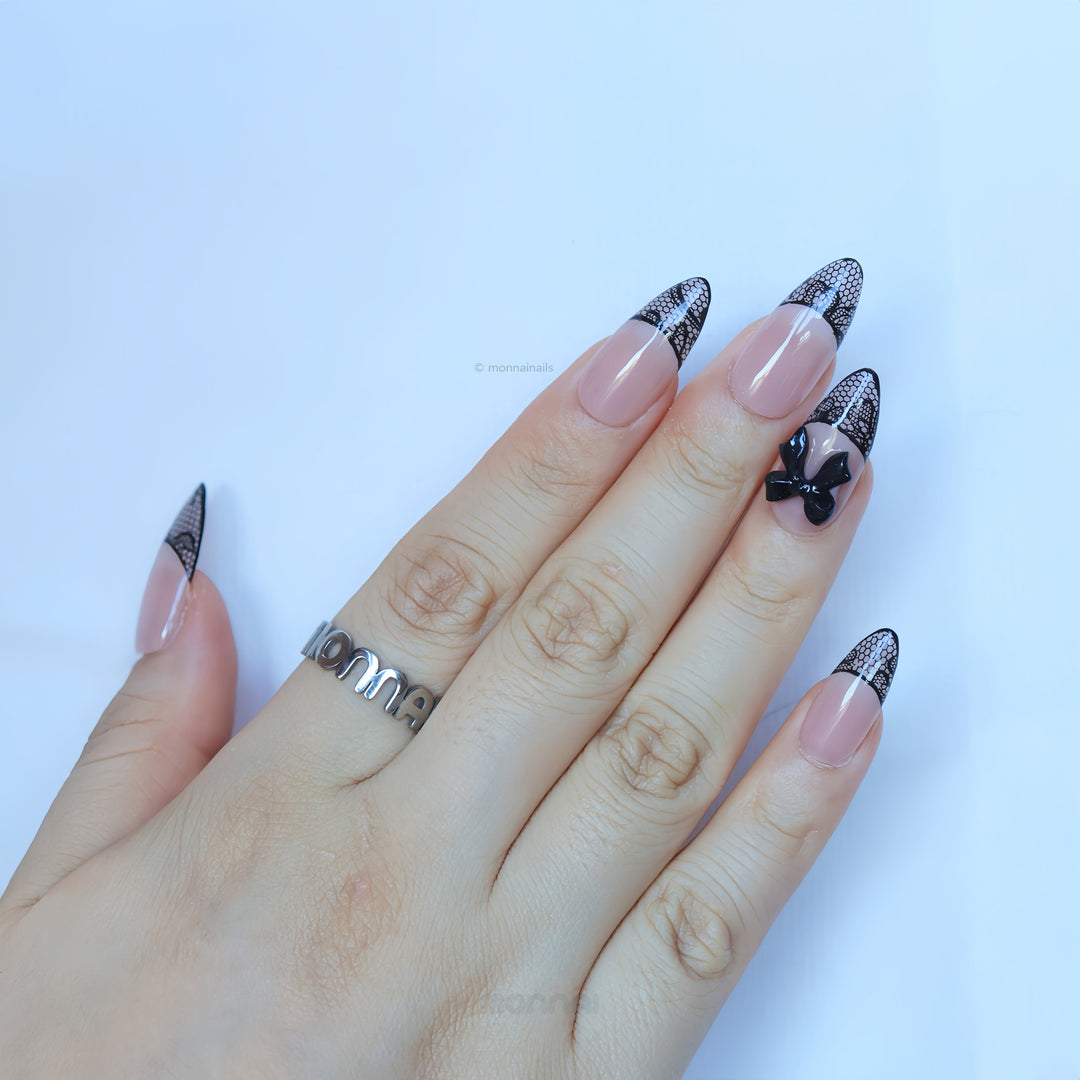 Pretty in Lace press-on nails worn on hand, sheer nude base with black lace French tips and a black bow accent.