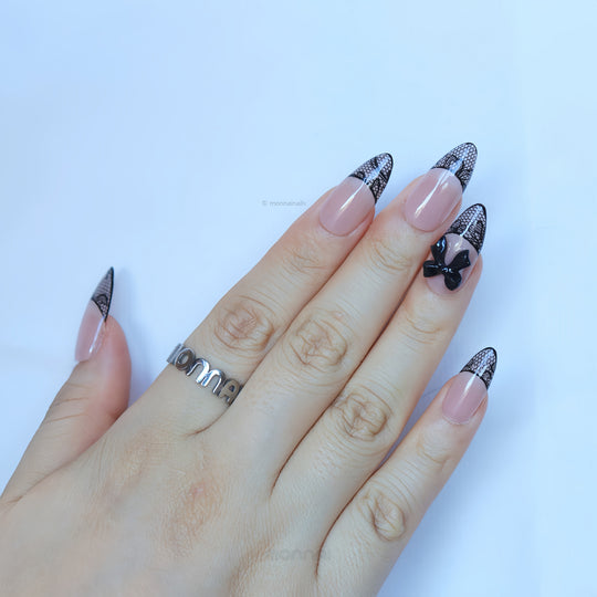 Pretty in Lace press-on nails worn on hand, sheer nude base with black lace French tips and a black bow accent.
