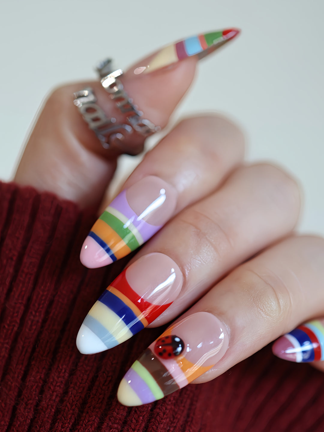 Rainbow Stripes press-on nails worn on hand with glossy nude base, colorful stripes, and a ladybug accent, playful