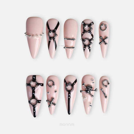 Rebellion press-on nails laid out in a set, with nude-pink tips, black harness linework, and silver O-ring hardware.