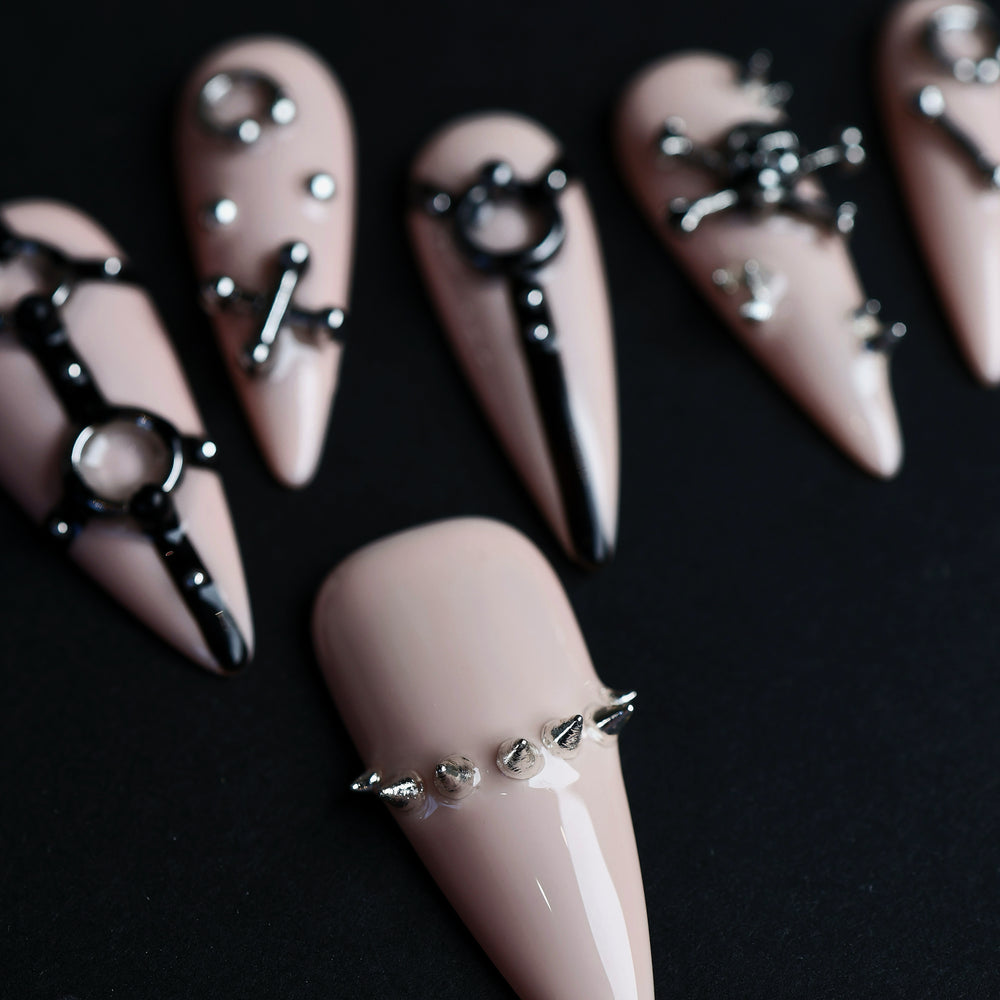 Rebellion press-on nails laid out in close-up, nude-pink with black harness linework and silver spikes.