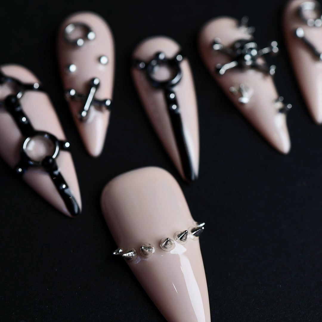 Rebellion press-on nails laid out in close-up, nude-pink with black harness linework and silver spikes.