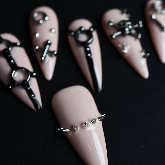 Rebellion press-on nails laid out in close-up, nude-pink with black harness linework and silver spikes.