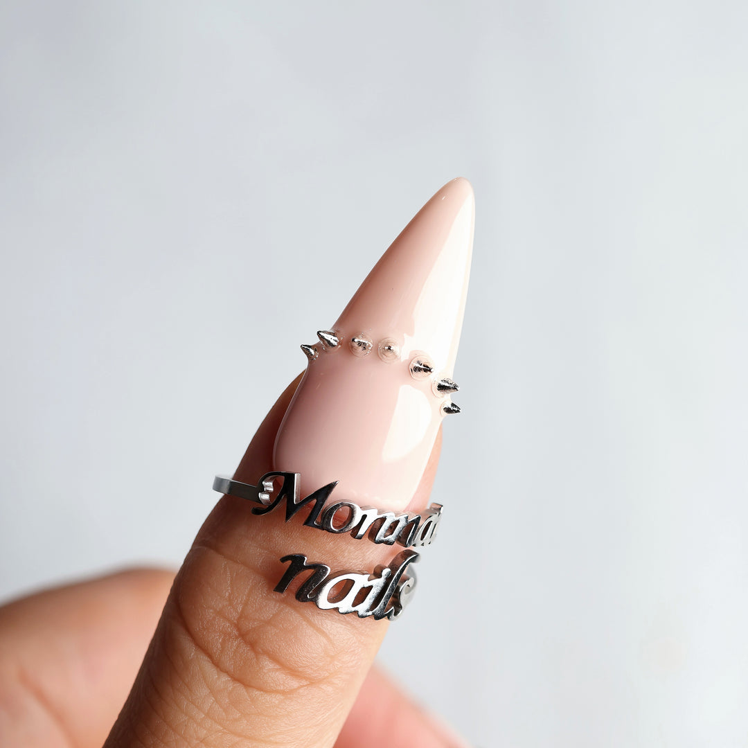 Rebellion press-on nails worn on hand, nude-pink stiletto nail with silver spikes and high-gloss finish.