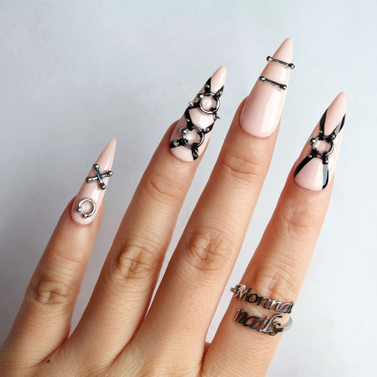 Rebellion press-on nails on hand, nude-pink with black harness linework and silver O-ring hardware.