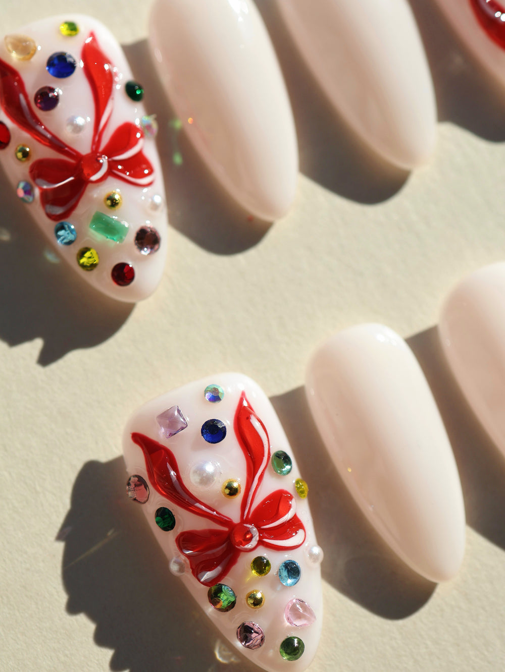 Red Bow press-on nails shown close-up, milky white almond nails with red 3D bows and multicolor rhinestones.