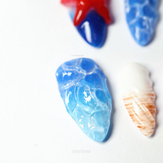 Seaside Fantasy press-on nails laid out in close-up, with blue water-ripple textures, a red starfish detail, and a shimmering silver base.