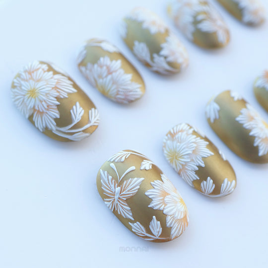 Silk &amp; Stitches press-on nails laid out, showing gold chrome with white floral embroidery details.