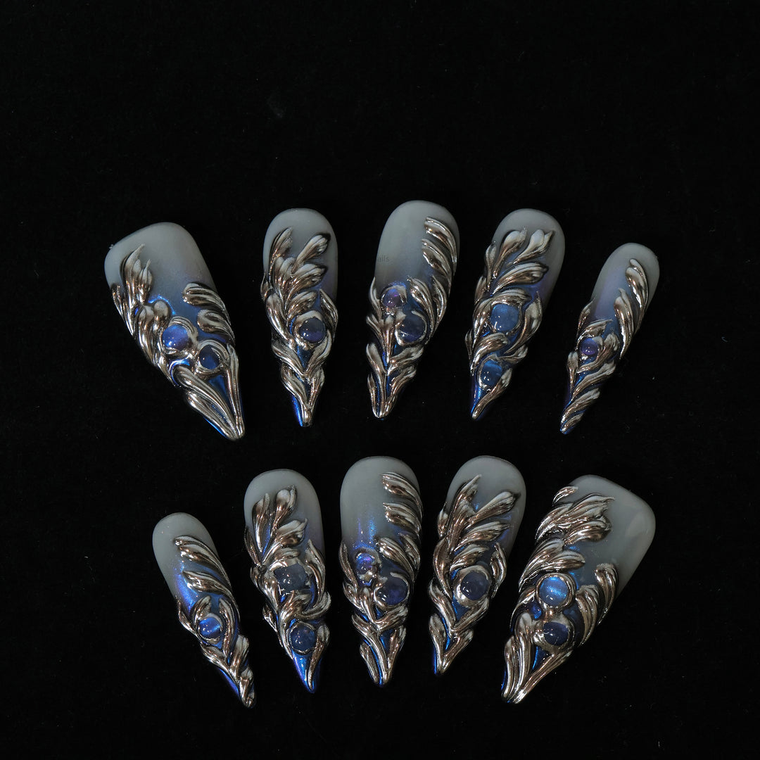 Silver Dreams press-on nails laid out in close-up, with icy gray bases, silver feather details, and blue iridescent accents.