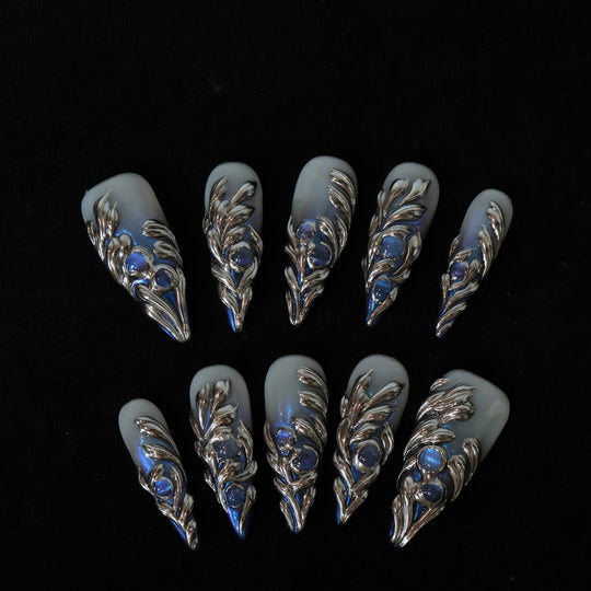 Silver Dreams press-on nails laid out in close-up, with icy gray bases, silver feather details, and blue iridescent accents.