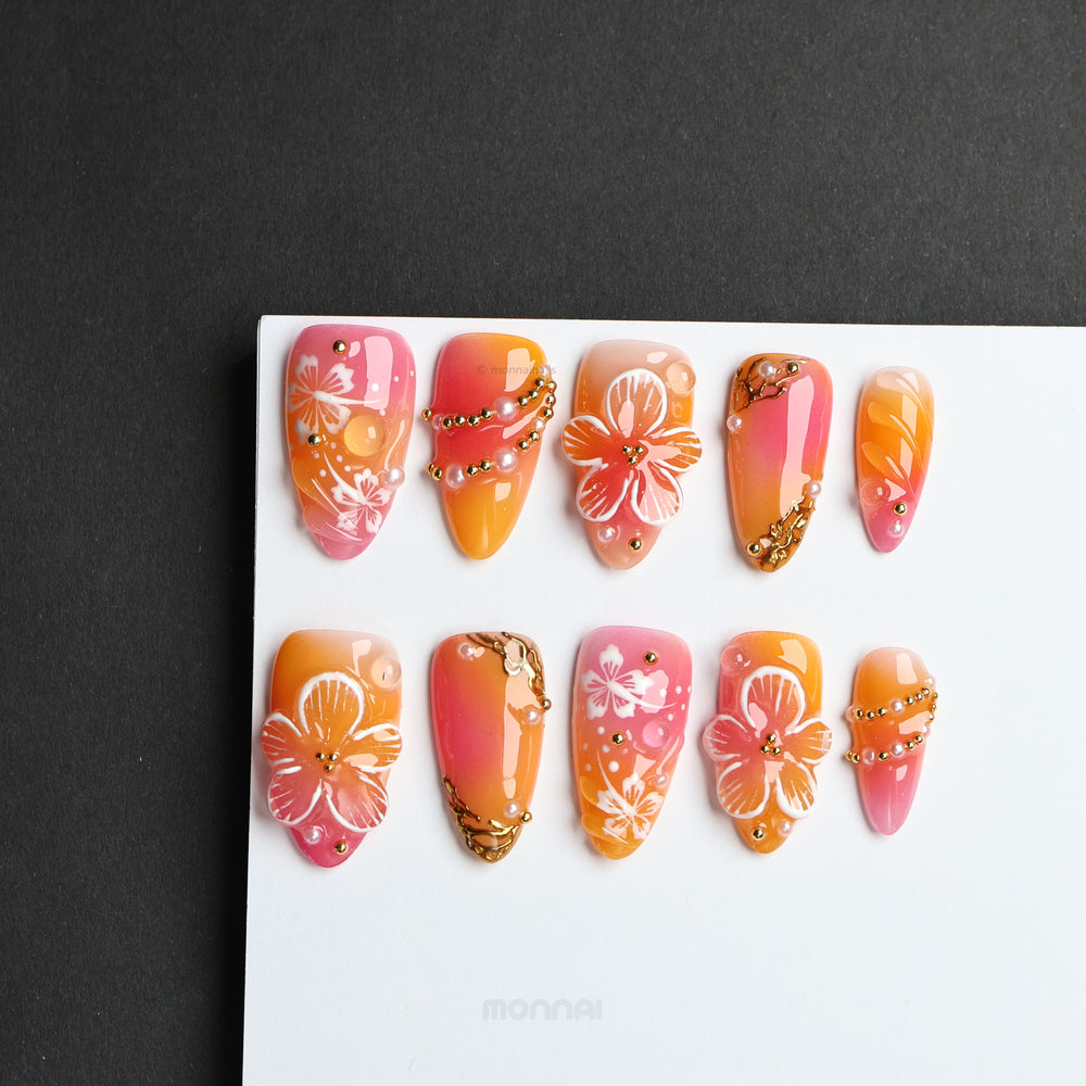 Sunset Hibiscus press-on nails laid out in warm pink and orange ombré with floral details and gold accents.