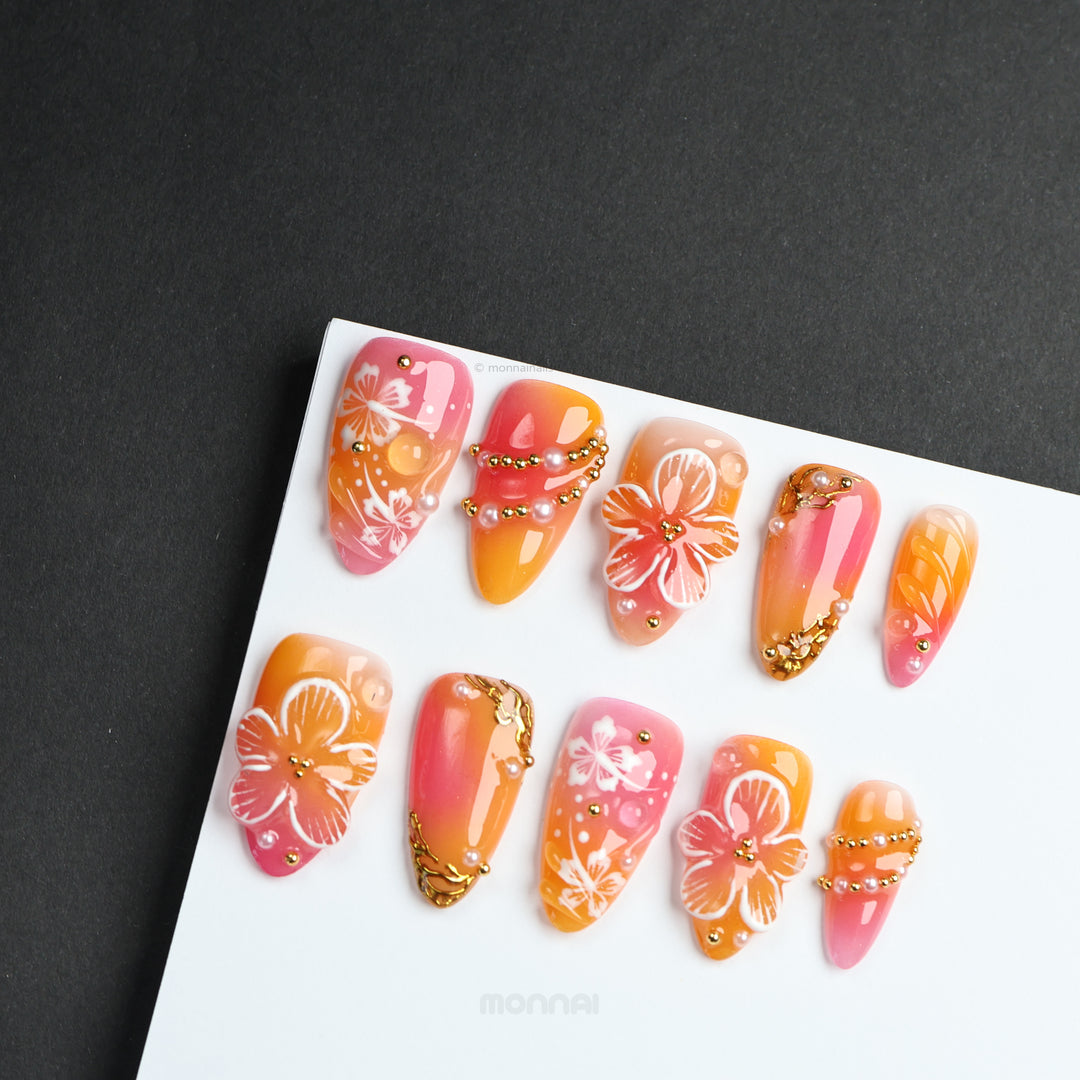Sunset Hibiscus press-on nails laid out in pink and orange ombré with hibiscus florals, gold accents, and crystals.