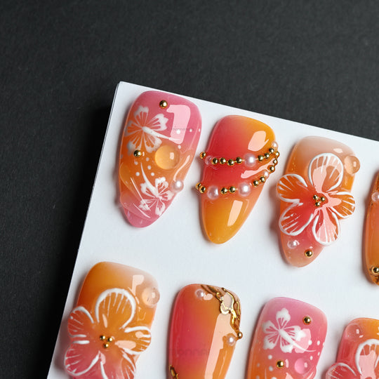 Sunset Hibiscus press-on nails laid out with glossy pink-orange ombré, floral details, and gold accents.