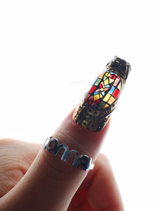 The Fifth Window press-on nails shown on a hand, with colorful stained-glass motifs in sculpted bronze frames.