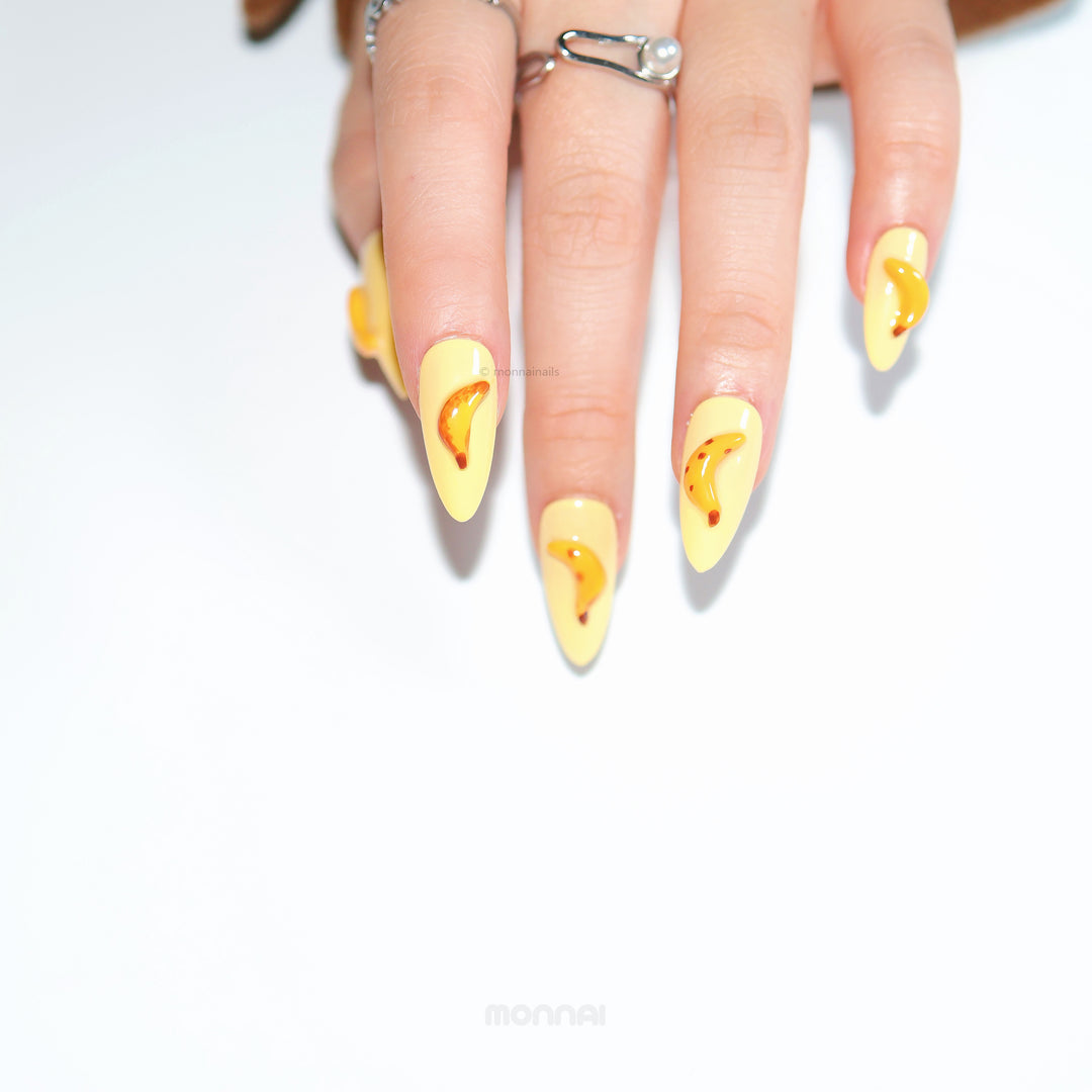 Tropical Banana press-on nails on hand, pale yellow with glossy banana accents in hyper-realistic pop art style.