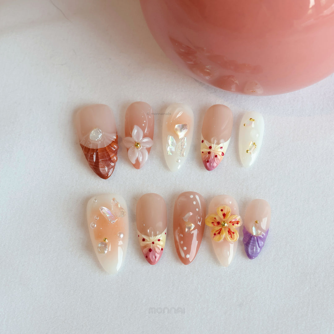 Winged Grace press-on nails laid out in soft peach and nude tones with butterfly and floral accents, pearls, and glossy translucent details.