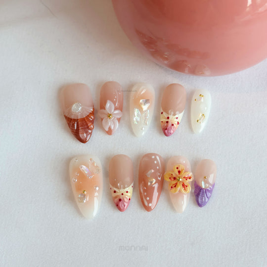 Winged Grace press-on nails laid out in soft peach and nude tones with butterfly and floral accents, pearls, and glossy translucent details.