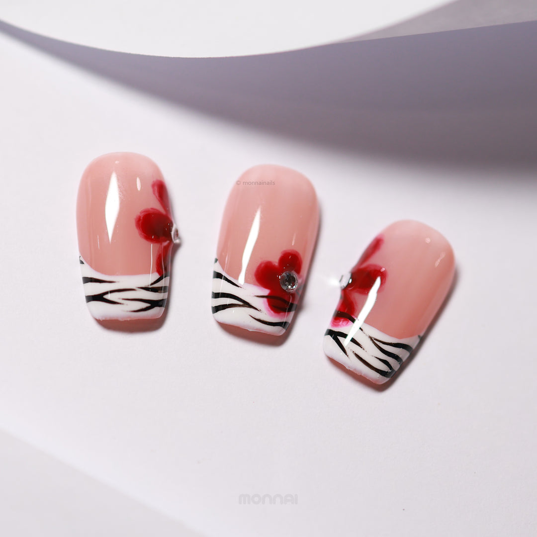 Zebra Blossom press-on nails laid out, showing nude bases, zebra French tips, and red floral accents with rhinestones.